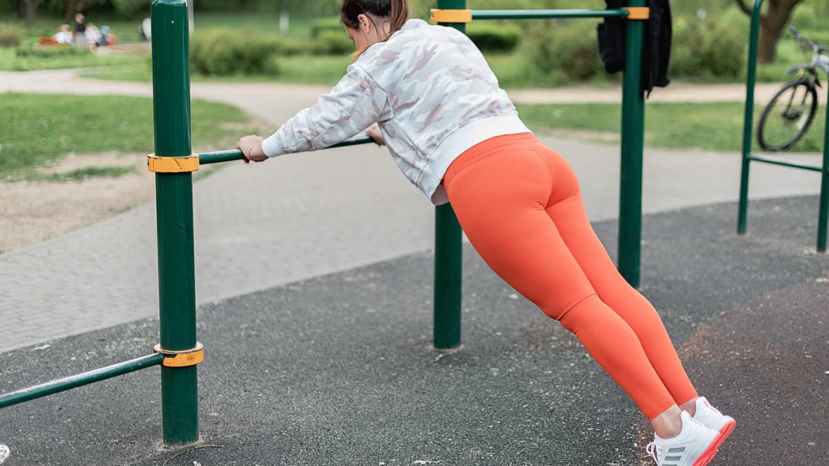 A woman in sportswear performs exercises on outdoor fitness equipment.