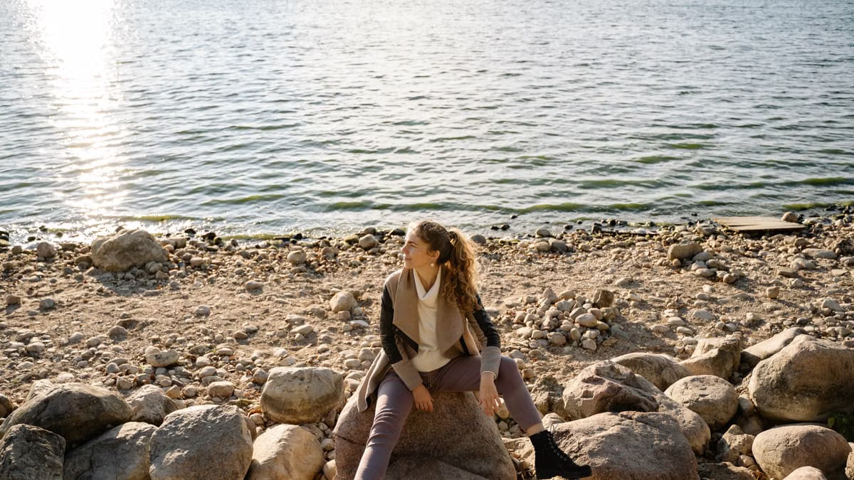 A woman sits on a rocky shoreline, gazing at the serene lake view during a sunny day.